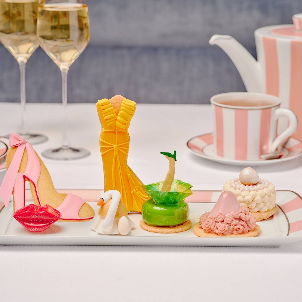 A stylish afternoon tea featuring fashion-inspired pastries, including a high heel, gown, and hat, served with champagne and striped china.
