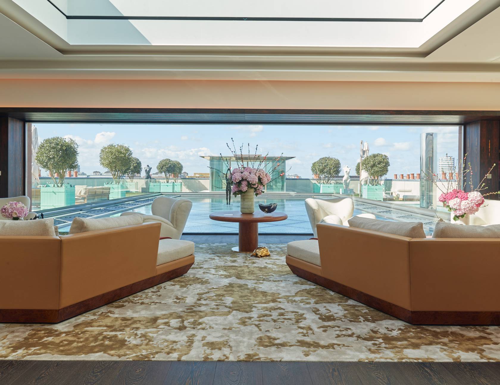 Luxurious rooftop penthouse lounge with skylight, plush seating, and floral arrangement, overlooking a serene private pool and manicured terrace against a blue-sky backdrop.
