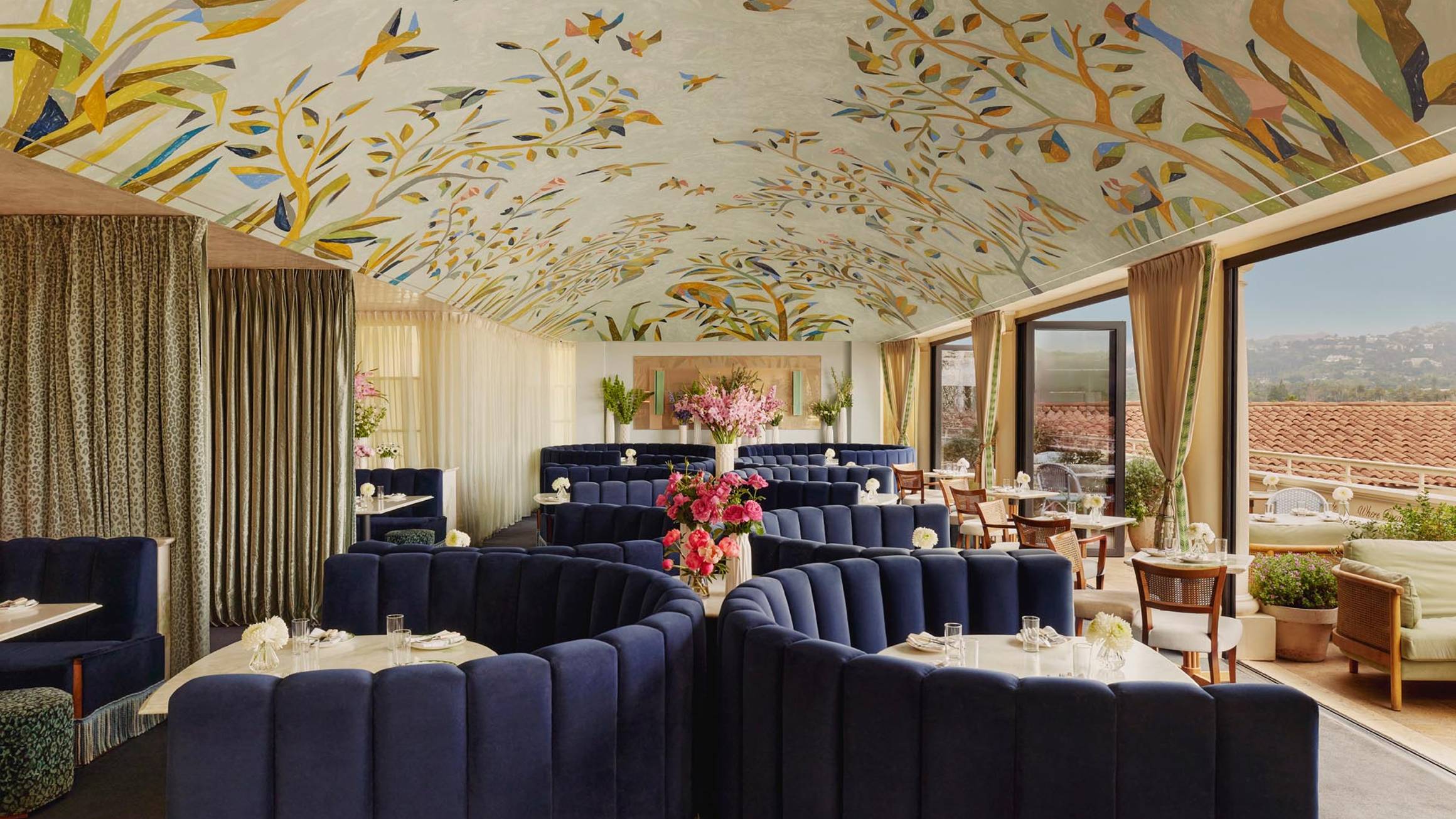 A sophisticated rooftop restaurant with plush navy booths, a hand-painted ceiling, and floor-to-ceiling windows overlooking Beverly Hills.