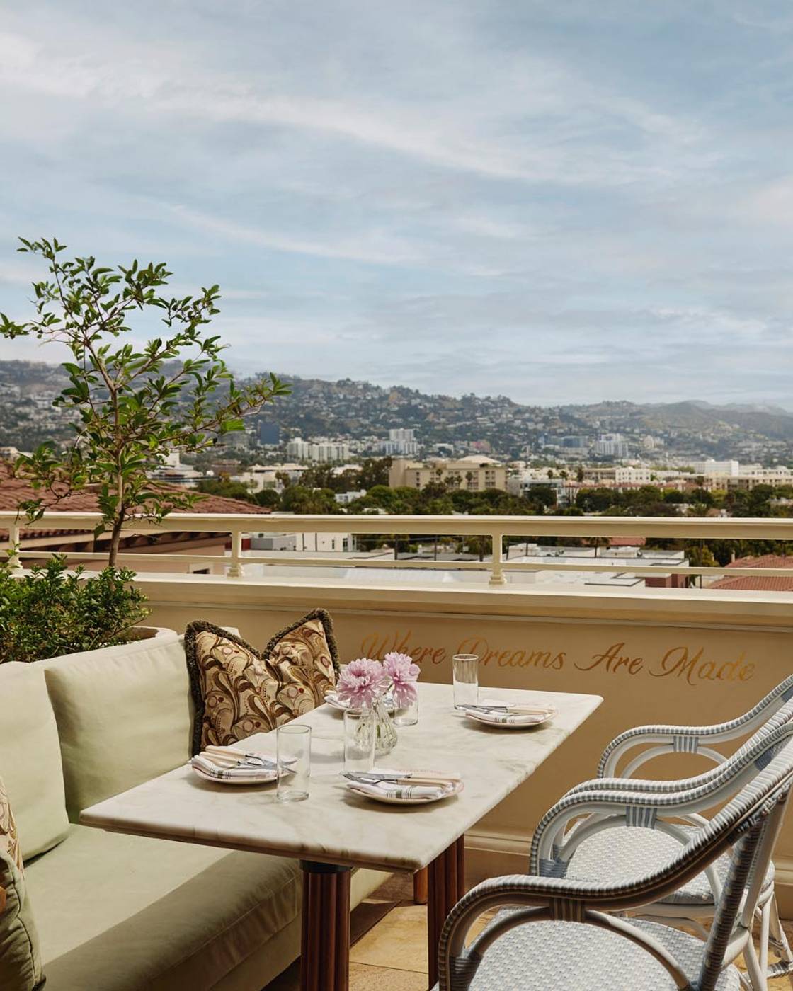 A sunlit terrace with elegant seating, marble tables, and panoramic city views, framed by lush greenery and soft furnishings.