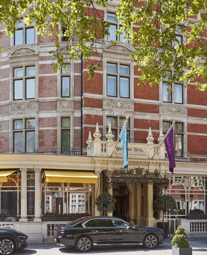 Exterior of The Connaught Hotel, featuring luxury cars parked outside, framed by elegant architecture and leafy trees overhead.