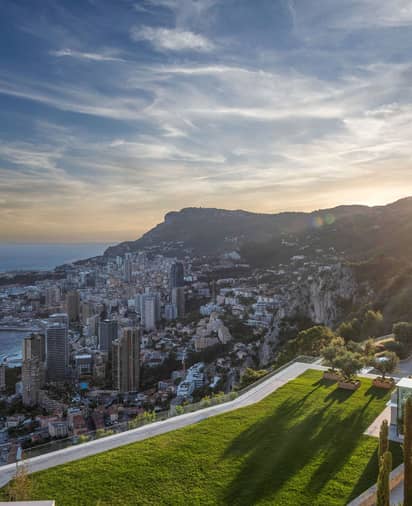 A breathtaking view of Monaco's coastline at sunset, with lush greenery, towering cliffs, and sparkling cityscape below.