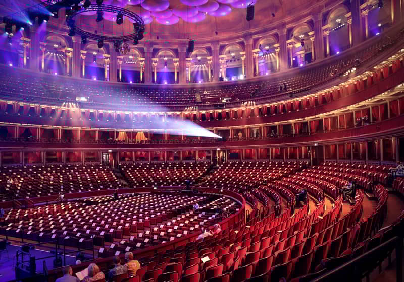 The grand interior of the Royal Albert Hall, featuring ornate balconies, glowing purple lighting, and rows of red velvet seats.
