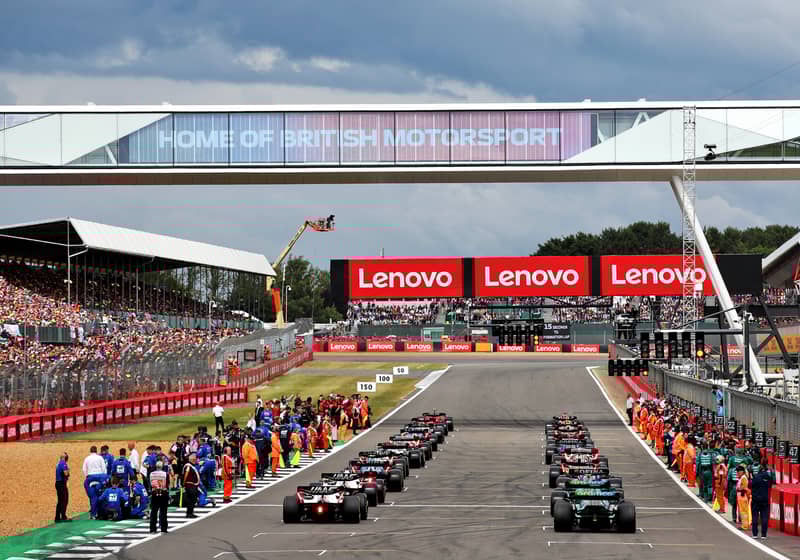 F1 cars lined up on the starting grid, surrounded by pit crews and a packed grandstand at Silverstone.