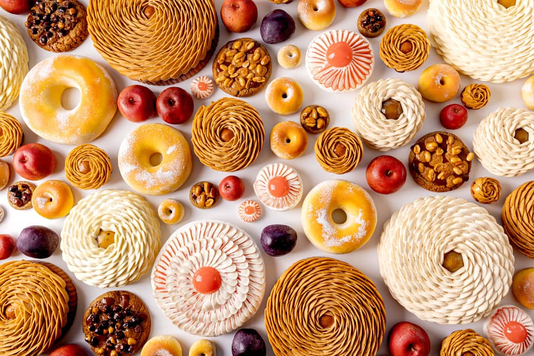 Overhead view of an exquisite assortment of pâtisserie creations in sculptural, fruit-inspired forms—featuring intricate spirals, glazes, and textures in soft ivory, caramel, and vivid red hues.