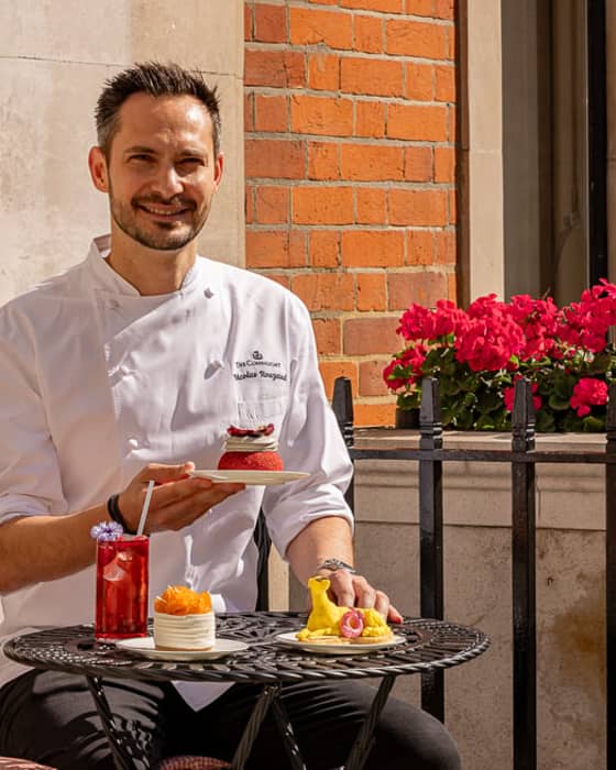A pastry chef in a white uniform seated at an outdoor café table, presenting elegant desserts with fresh fruit and garnishes.
