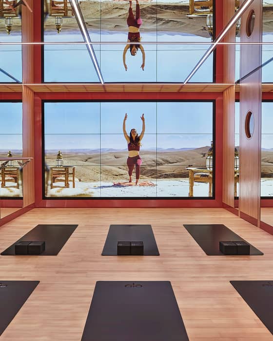 A tranquil yoga room with mirrored walls, wooden flooring, and a digital projection of a desert wellness retreat.