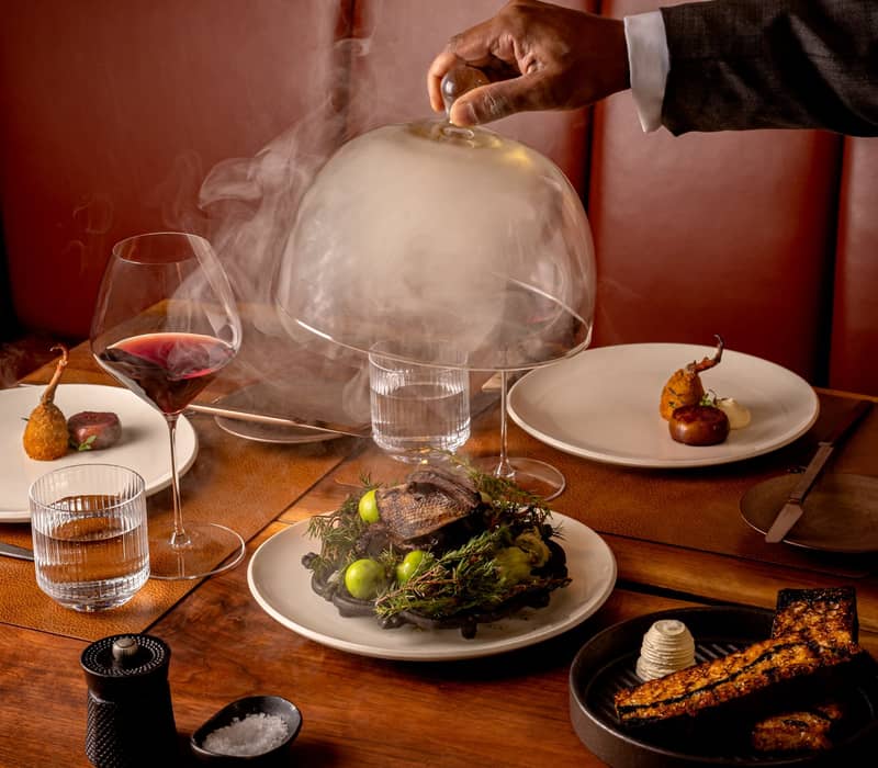 Luxurious dining setup featuring a smoked entrée unveiled under a glass cloche, paired with fine wine and artistic sides.