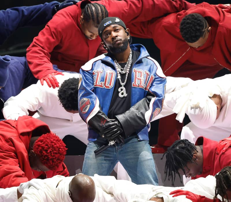 endrick Lamar on stage in a blue “GLORY” jacket, surrounded by dancers in red, white, and blue outfits.