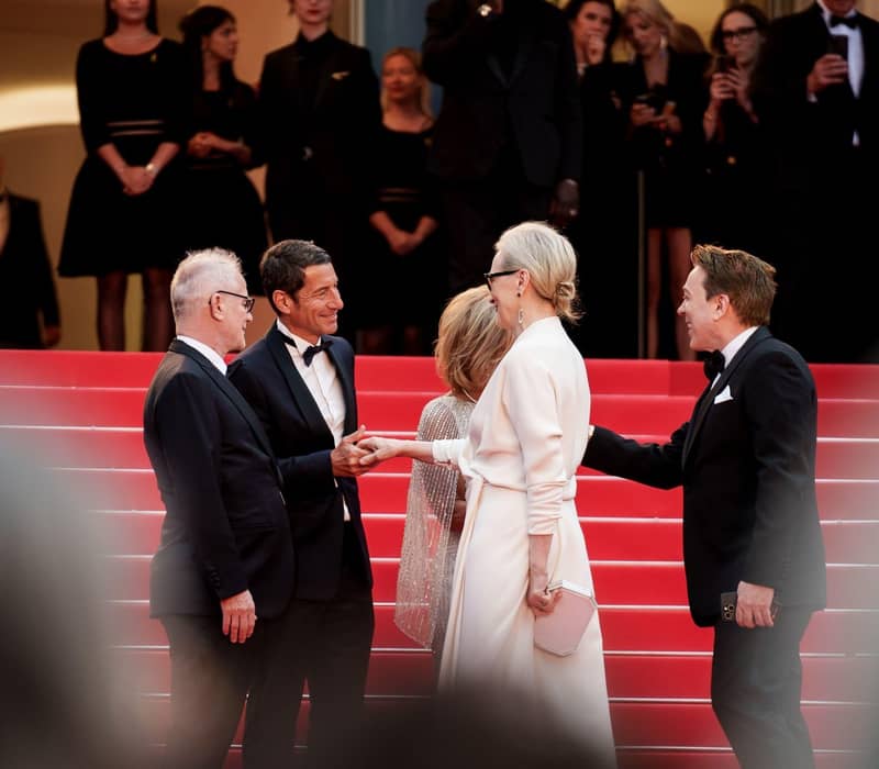 Celebrities greet each other on red carpet stairs, surrounded by elegantly dressed attendees at Cannes Film Festival