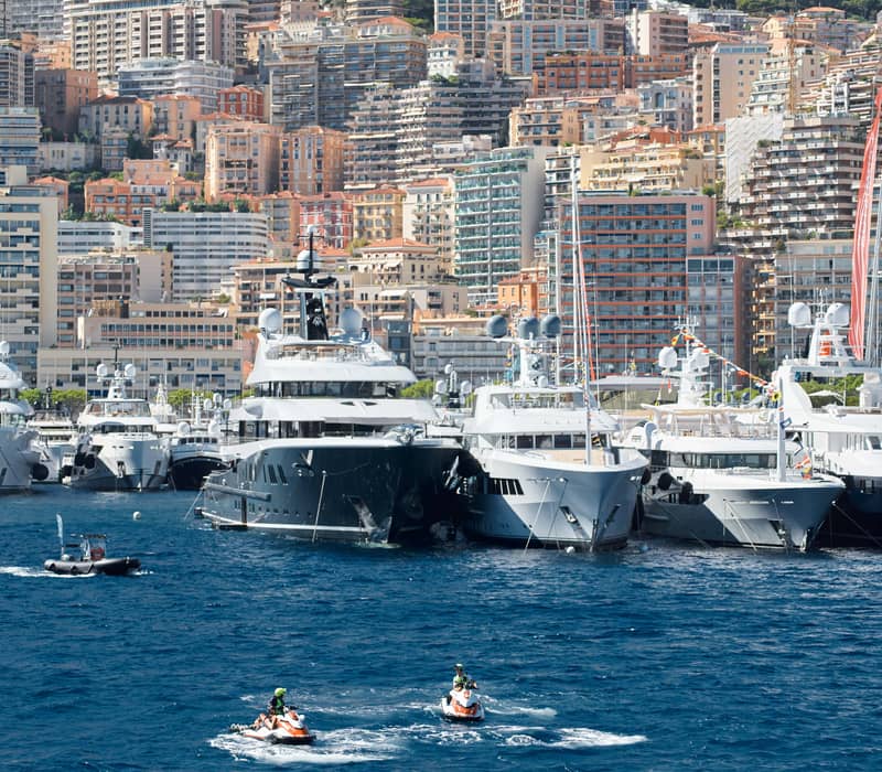 A dazzling display of superyachts moored against Monaco’s hillside skyline, with jet skis gliding across the deep blue water.