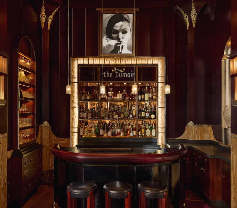 Decadent cocktail bar with Art Deco details, rich burgundy tones, illuminated shelving, and an intimate ambiance framed by soft lighting