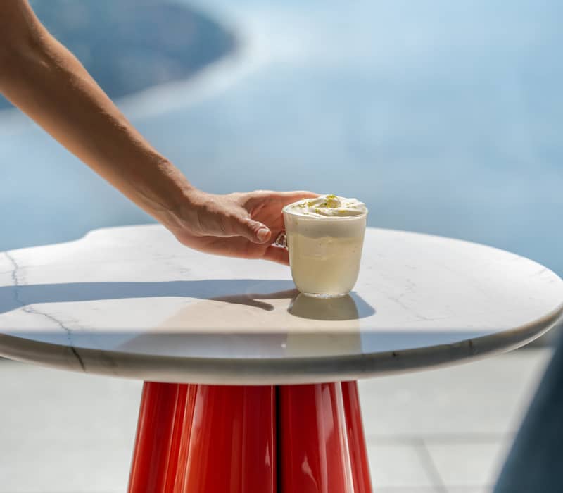 Minimalist drink service with a hand placing a cocktail on a marble table overlooking an azure ocean view.
