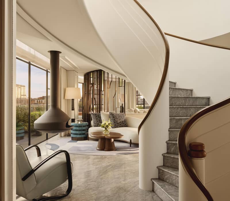Striking spiral staircase in Claridge’s The Residence opens to a bright lounge with sculptural furniture, round coffee table, and suspended fireplace.
