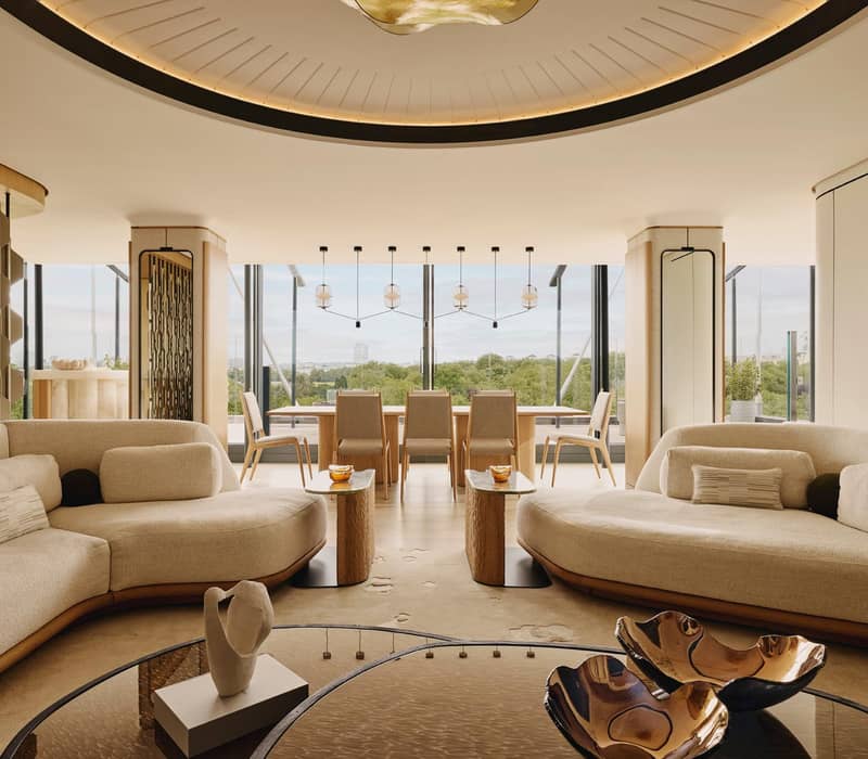 Luxurious penthouse with curved sofas, modern decor, dining area, and floor-to-ceiling windows overlooking lush greenery