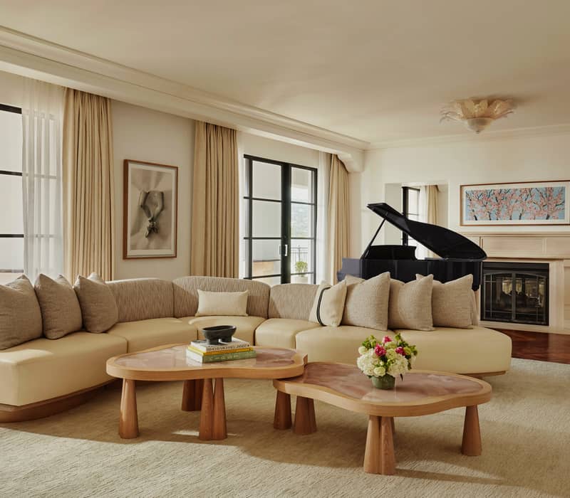 Elegant living room with cream sectional sofa, wood coffee tables, grand piano, and natural light from large windows