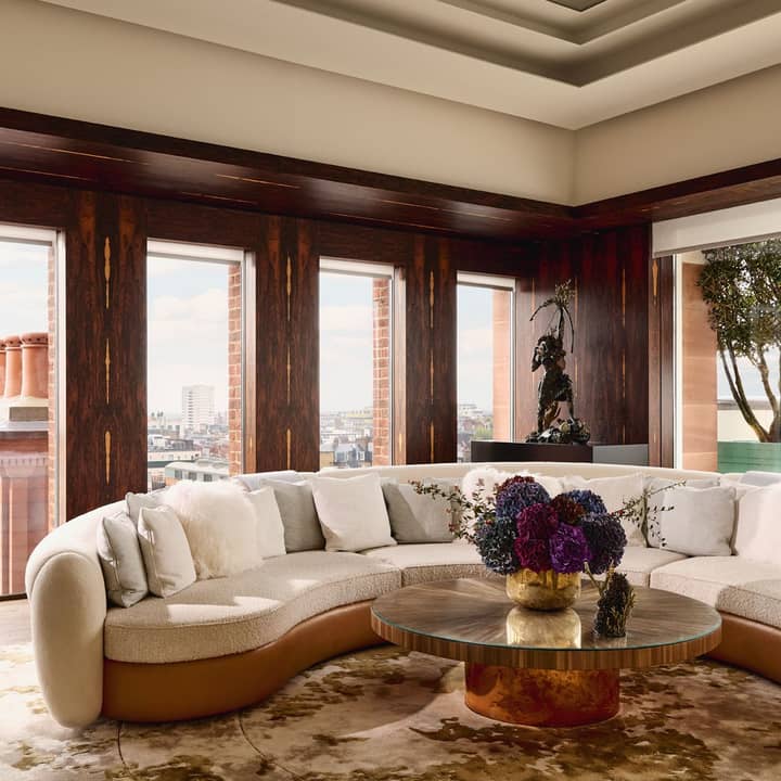 A refined lounge area featuring a curved cream sofa, dark wood paneling, panoramic city views, and a statement coffee table with floral arrangements.