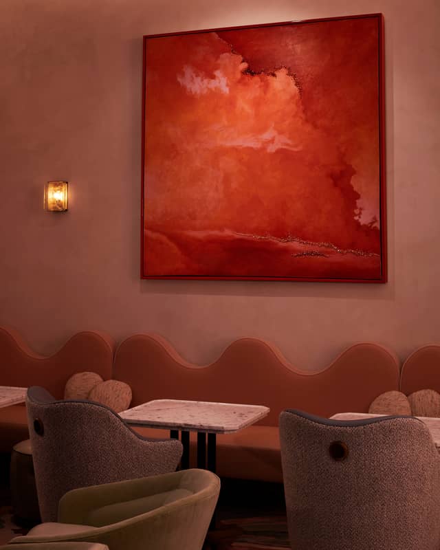 Warm-toned room featuring curved banquettes, marble tables, and a bold abstract painting in rich red hues