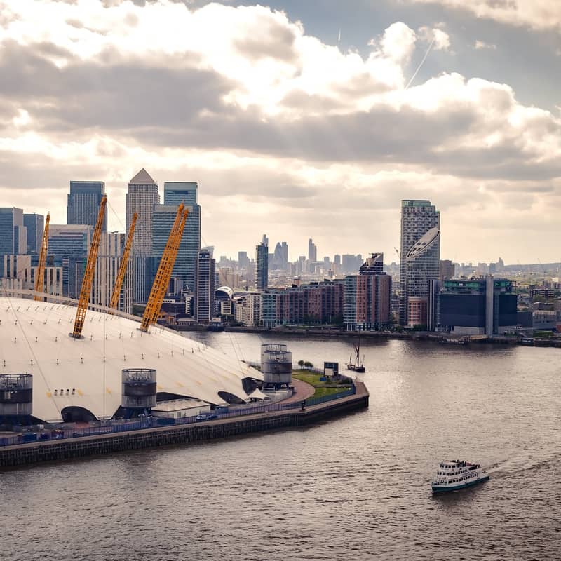 Panoramic view of The O2 and Canary Wharf skyline, framed by the Thames, under a cloudy yet luminous sky.