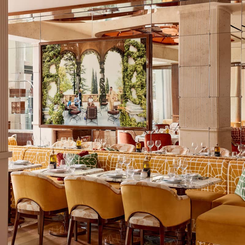 A stylish dining space featuring mustard-yellow seating, mirrored walls, and an ornate framed artwork evoking a Mediterranean atmosphere.