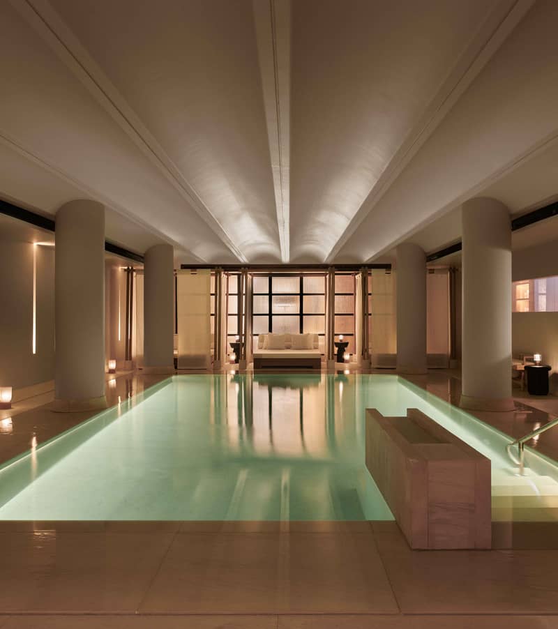 A serene indoor swimming pool with soft lighting, elegant columns, and a luxurious daybed at the far end.