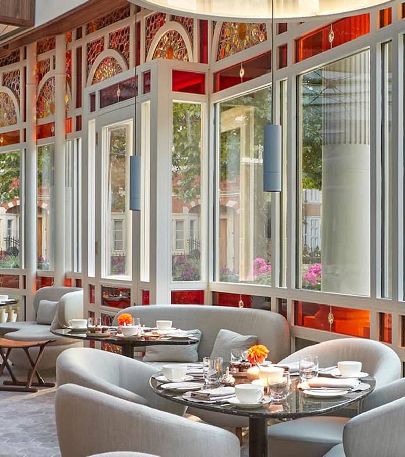 A sunlit restaurant with plush seating, glass walls, and vibrant decor overlooking a picturesque city street.