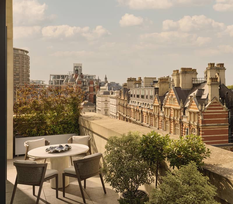 View from a private terrace featuring a round dining table with four cushioned chairs, surrounded by greenery, overlooking red-brick Victorian rooftops and treetops under a bright, partly cloudy sky.