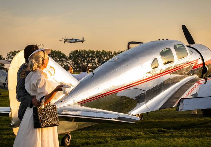 A stylish couple in vintage attire admiring a polished classic aircraft at an exclusive motorsport and aviation event.