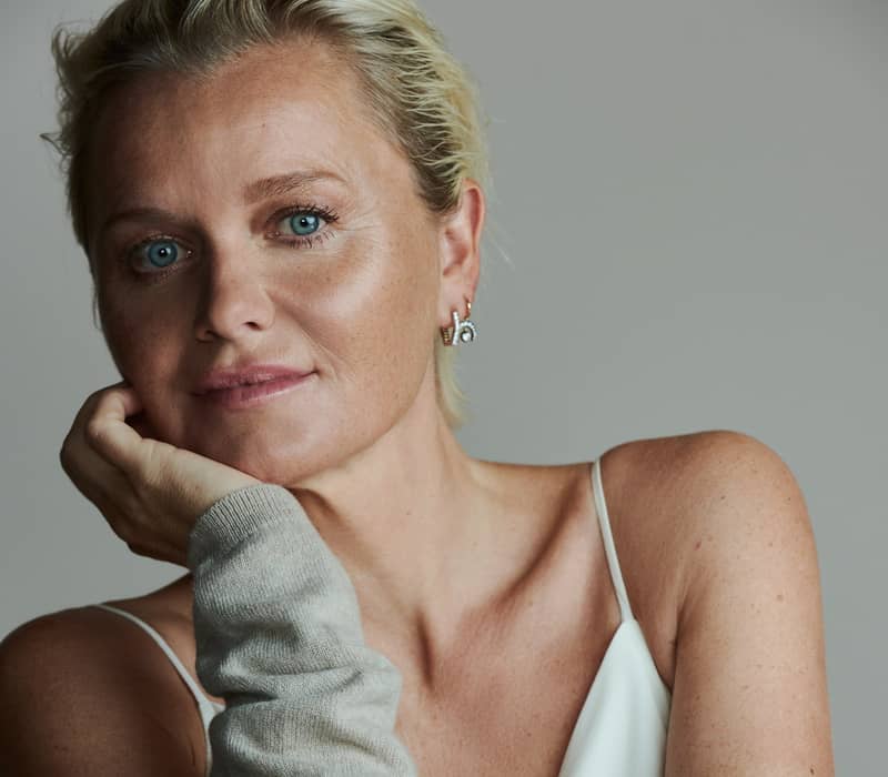 Dr. Barbara Sturm, gracefully posed with her chin resting on her hand, radiates elegance and natural beauty in minimalist attire.