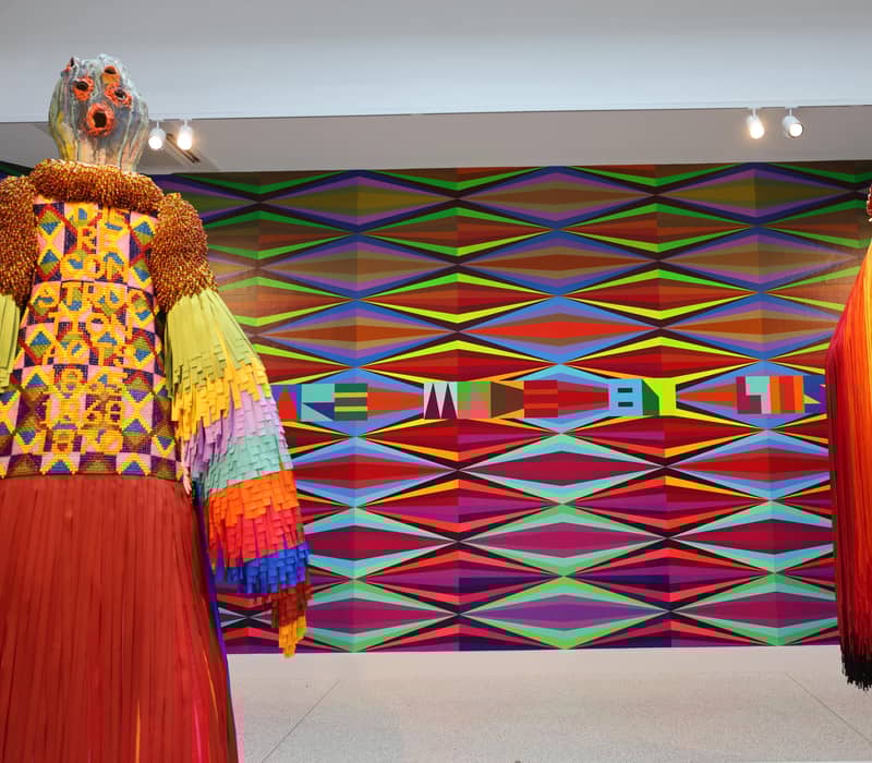 Two vibrant textile sculptures face a colorful geometric mural reading “THE SPACE MADE BY US.”