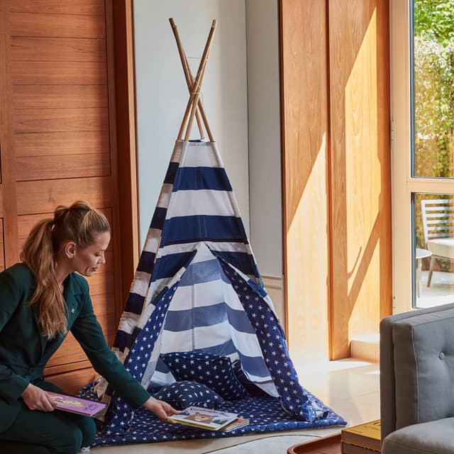 A woman in a dark green suit arranges children's books beside a small blue and white striped play tent with star-patterned lining, set in a sunlit room with wooden panels and large windows.