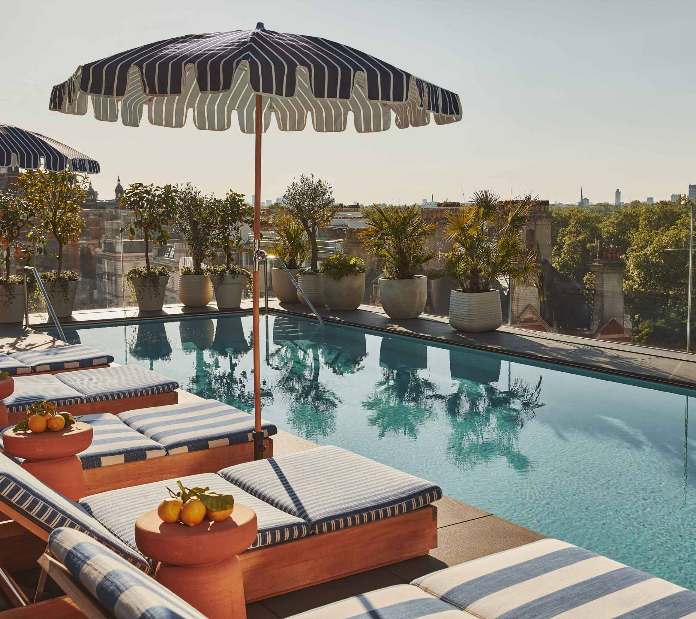 Lounge chairs and striped umbrellas beside a rooftop pool, with potted plants and a city view.