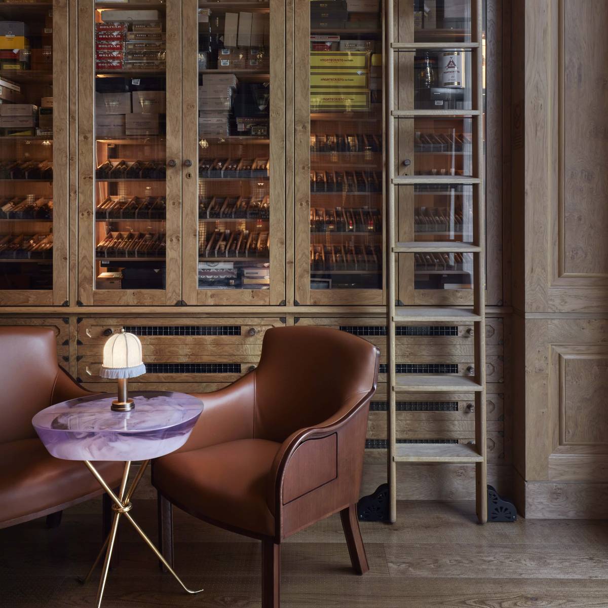 Elegant cigar humidor with wooden cabinetry, a sliding ladder, leather armchairs, and a marble-topped table with ambient lighting