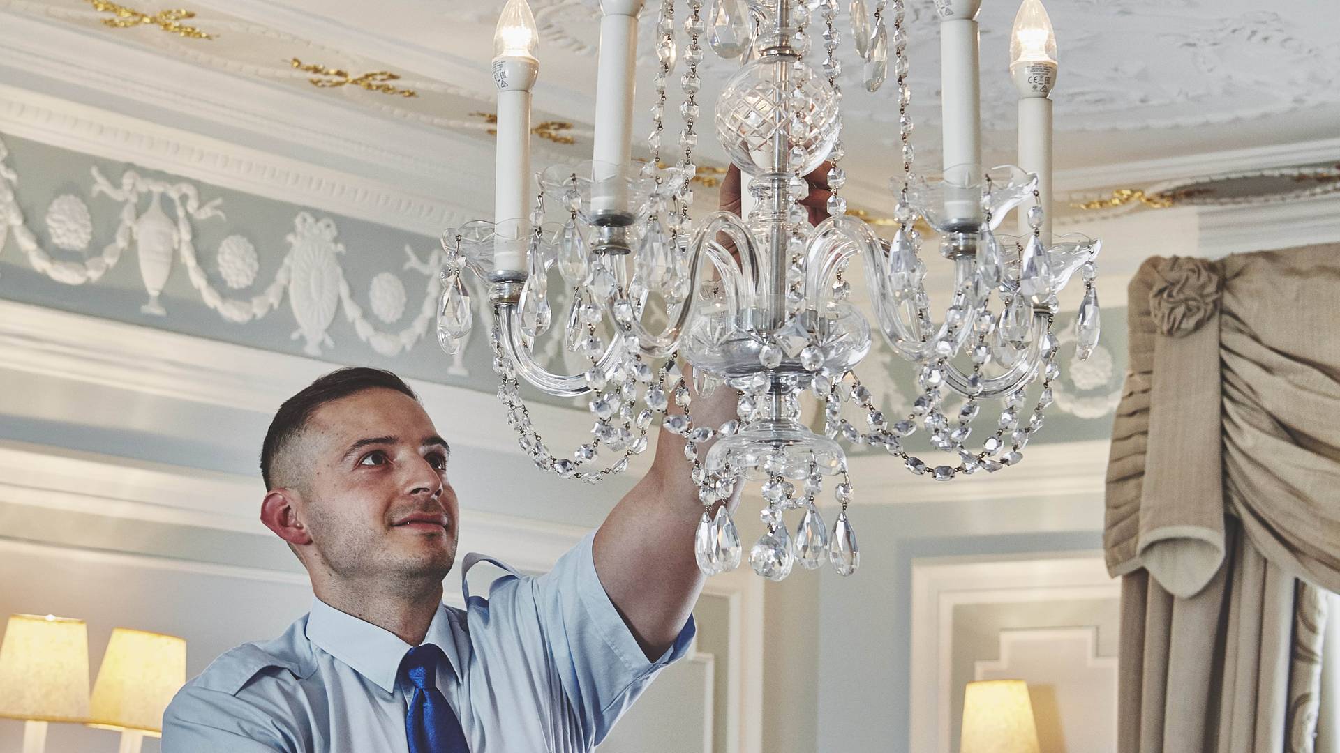 A technician in a blue uniform carefully adjusts a crystal chandelier in an opulent room with intricate gold ceiling details.