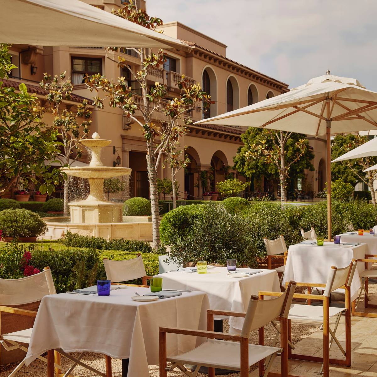 An elegant outdoor terrace with white linen-covered tables, wooden chairs, and umbrellas, surrounded by lush greenery and a central fountain.