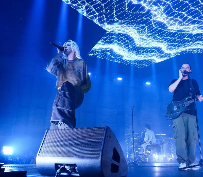 Linkin Park members perform under vibrant blue lights, with digital visuals overhead and crowd visible in the background.