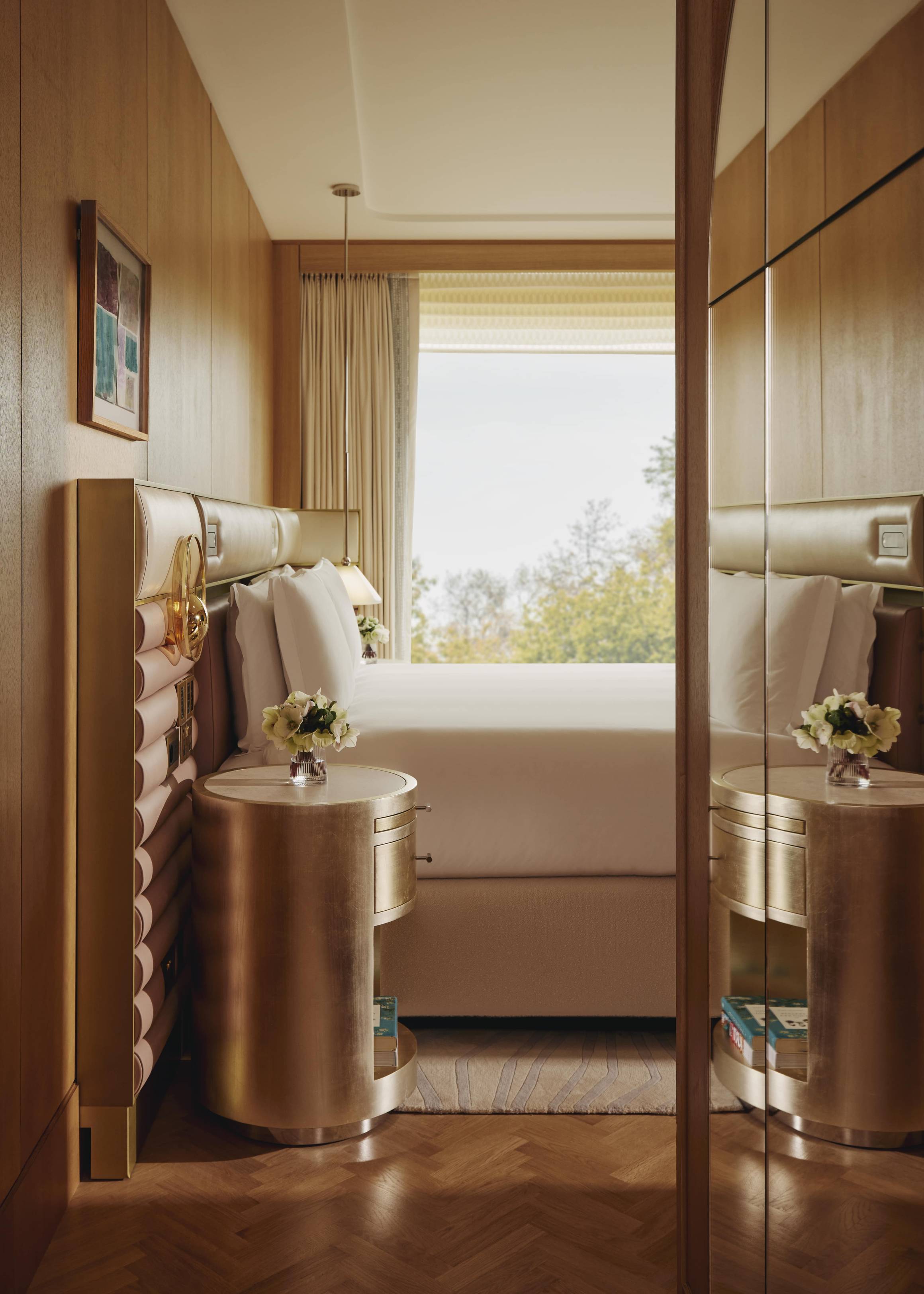 Luxurious bedroom with gold-trimmed headboard, mirrored panels, and large window overlooking treetops, evoking calm sophistication.