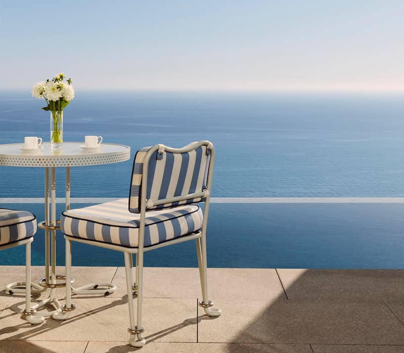 Bistro table with striped chairs and flowers overlooks calm ocean under clear blue sky, with stone terrace in foreground.
