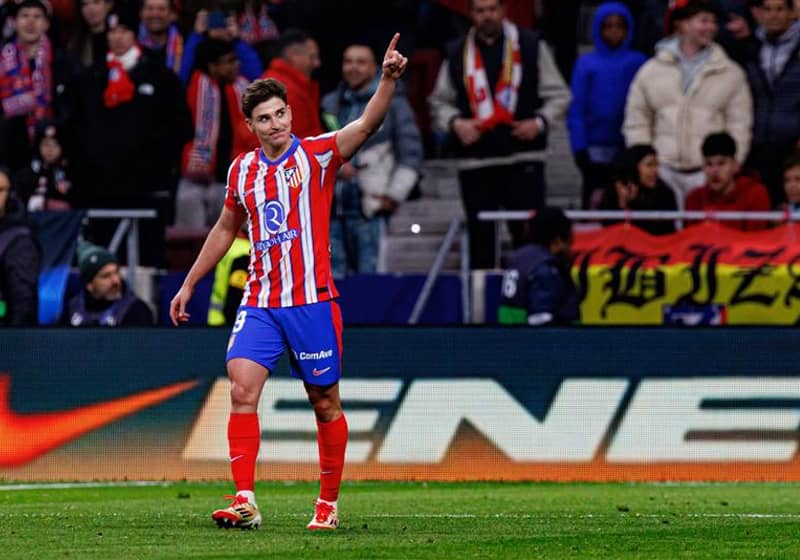 Soccer player in red and white striped kit celebrates a goal, pointing upward on field with cheering crowd in background.