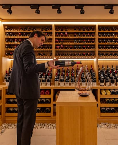Sommelier pouring wine into a decanter in a beautifully lit, wooden wine cellar stocked with premium bottles.