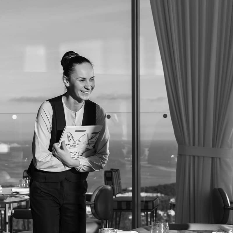 A smiling waitress holds a menu in a sunlit restaurant with panoramic views, exuding warmth and elegance.