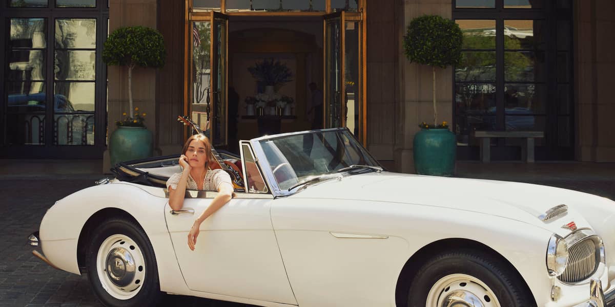 A woman reclines in a vintage white convertible, exuding effortless elegance in front of a grand entrance with classic architecture.