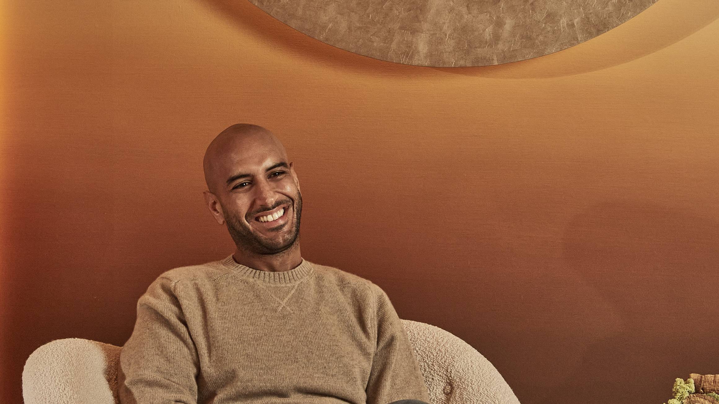 A man with a warm smile, dressed casually in a beige sweater and jeans, sits on a textured chair in front of a soft gradient backdrop with a circular decorative element.