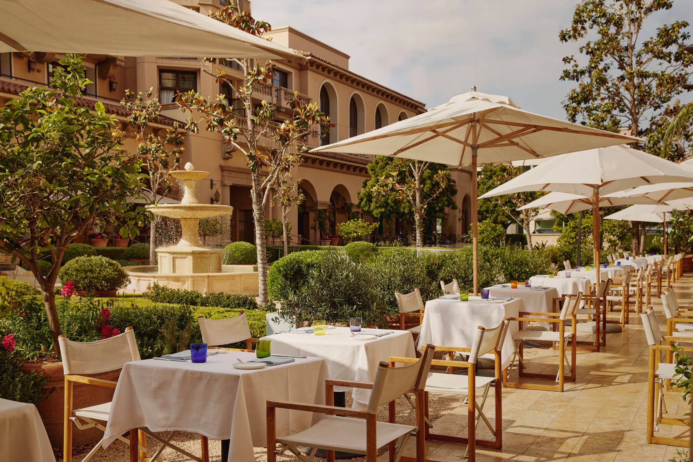 Elegant outdoor terrace dining with white tablecloths, large parasols, and a central stone fountain in manicured gardens.