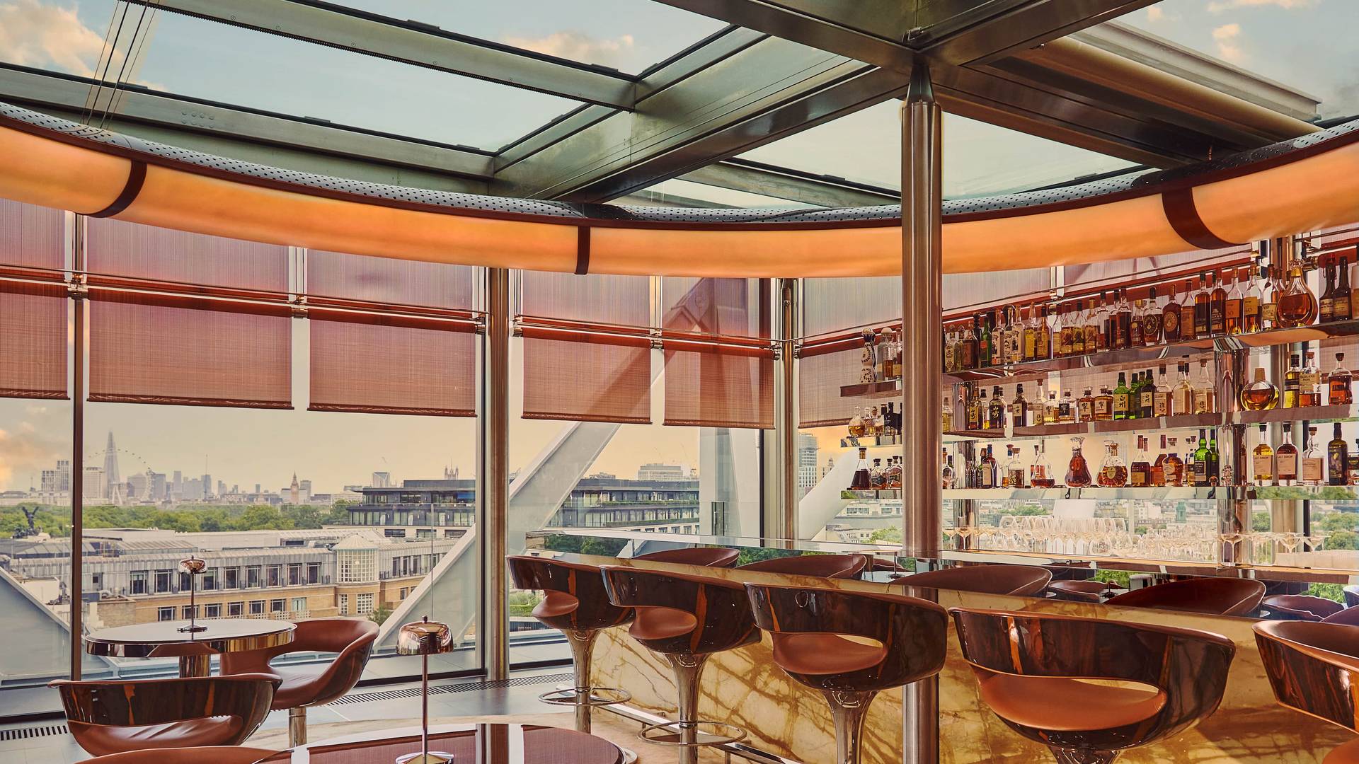 Rooftop bar with marble counter, leather stools, and round tables set beside floor-to-ceiling windows offering panoramic city views at sunset.