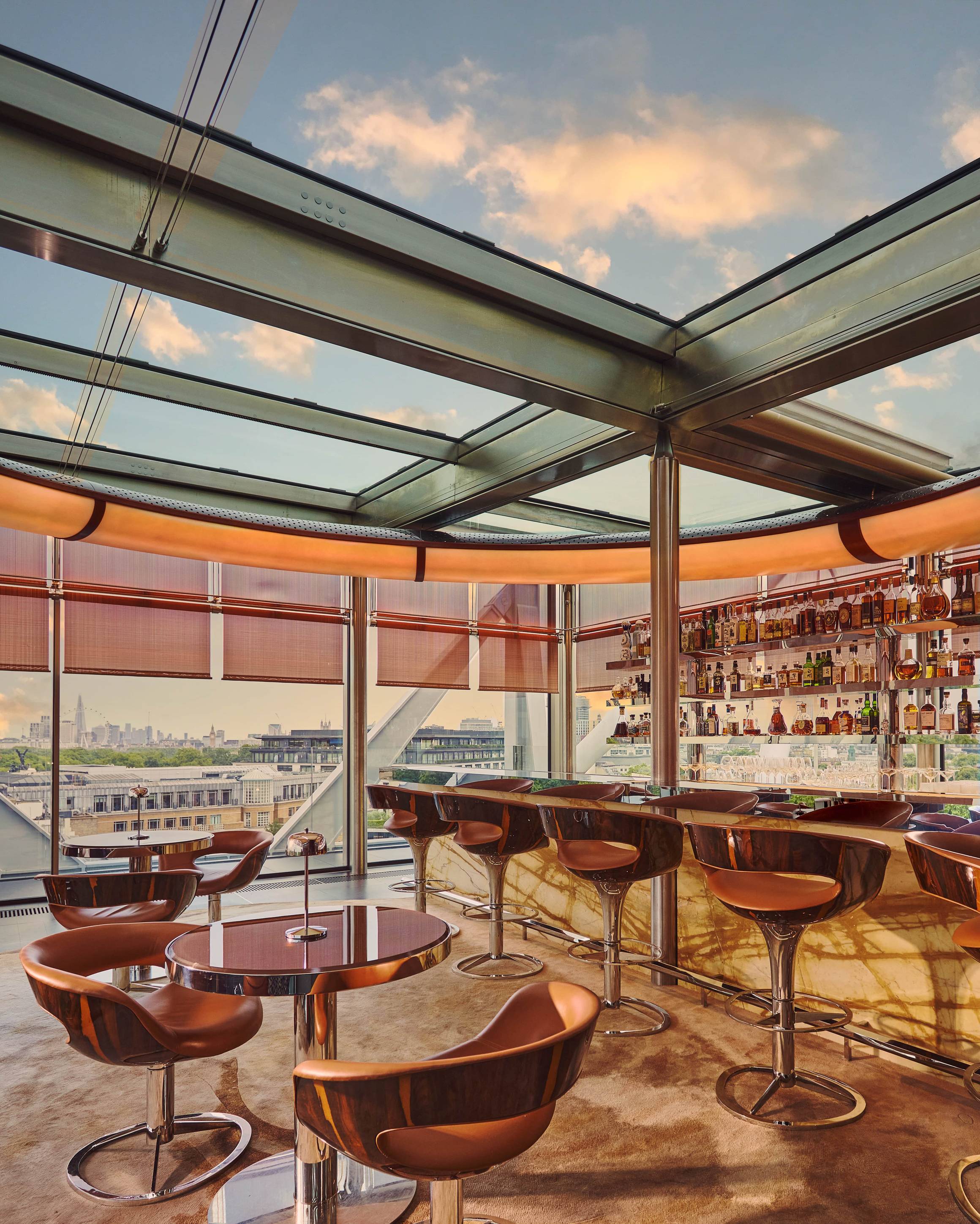 Rooftop bar with marble counter, leather stools, and round tables set beside floor-to-ceiling windows offering panoramic city views at sunset.