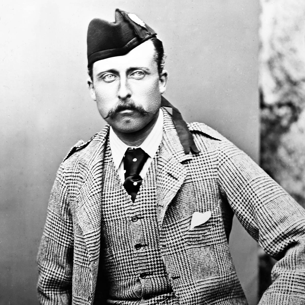 Man in traditional Scottish attire with mustache, checkered suit, and feathered hat, seated against plain background.