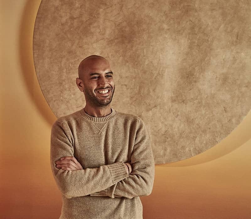Dr. Mark at Surrenne stands confidently, smiling, in front of a large, textured circle art piece on a warm-toned wall