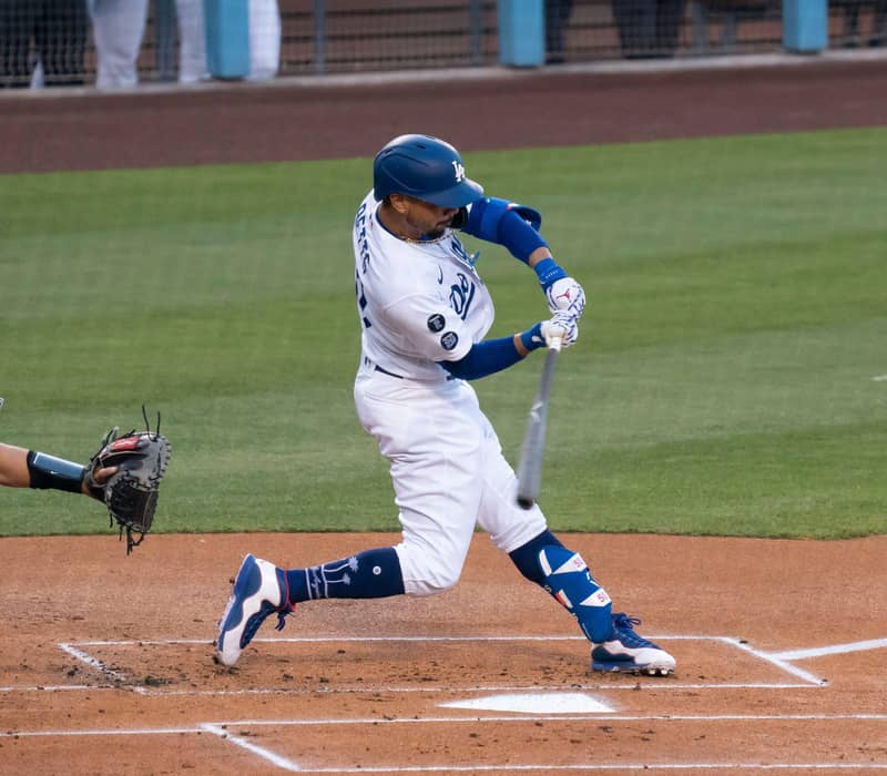 Baseball player in Dodgers uniform swings bat at incoming pitch during game, with catcher ready behind home plate.