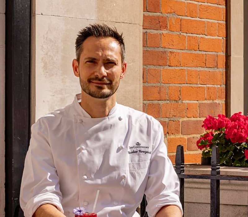 Chef in a white coat seated outdoors, presenting an array of vibrant pastries and a refreshing drink on a café table